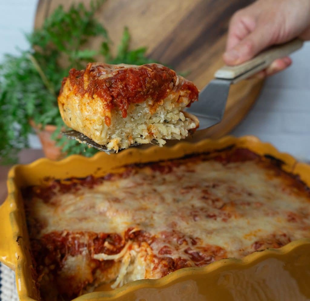 spatula lifting a piece of spaghetti pie out of the dish of spaghetti pie. dish is a gold color, there is a plant and a wood cutting board in the background, white tile backsplash
