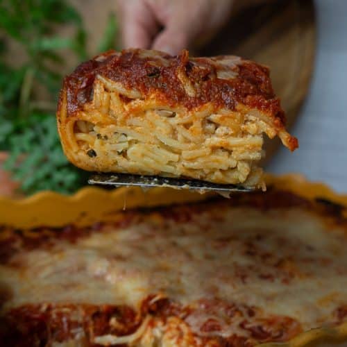square slice of spaghetti pie on a spatula being held by a hand over the dish of spaghetti pie