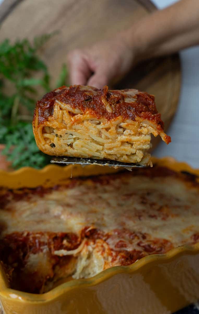 square slice of spaghetti pie on a spatula being held by a hand over the dish of spaghetti pie