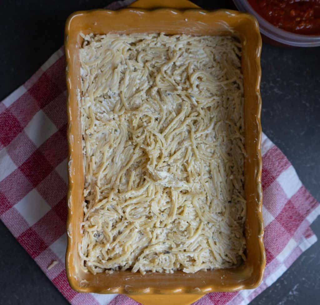 spaghetti and cheese mixture pressed evenly into the bottom of a 9x7 gold casserole dish. dish is setting on a red and white kitchen towel the countertop is black