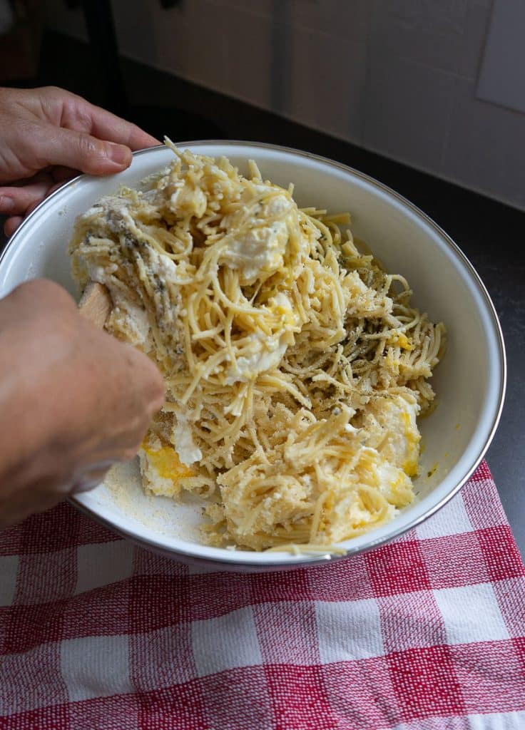 mixing the spaghetti and cheese mixture together in white bowl over a red and white checked kitchen towel