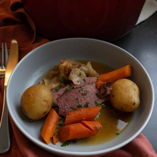 corned beef sliced and served on a gray plate with carrots, potatoes, and cabbage with pan juices