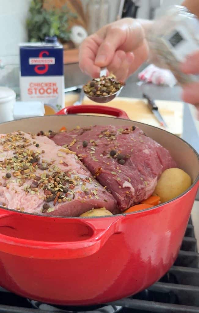 corned beef in pot with vegetables on stovetop hand is sprinkling spices over meat