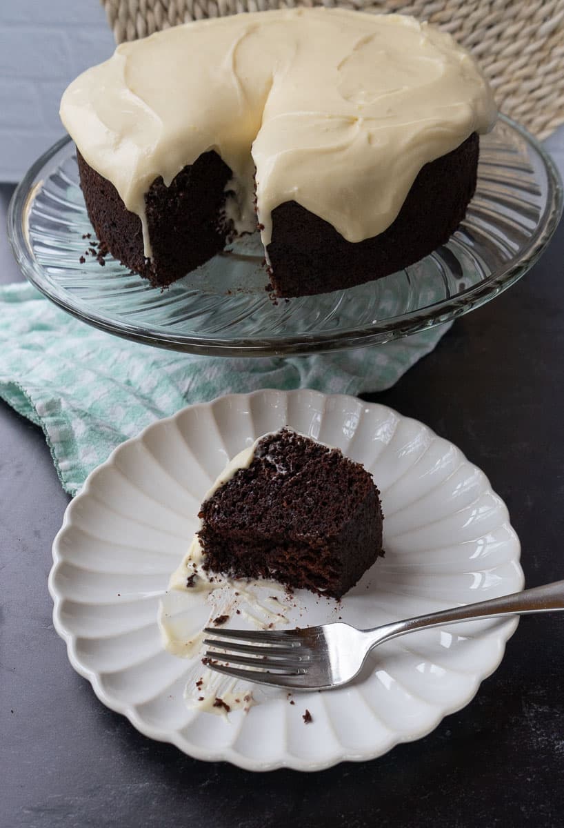 chocolate cake on a glass plate stand with slice of guinness stout chocolate cake on a white plate with a green and white gingham napkin under the plate. fork is on plate
