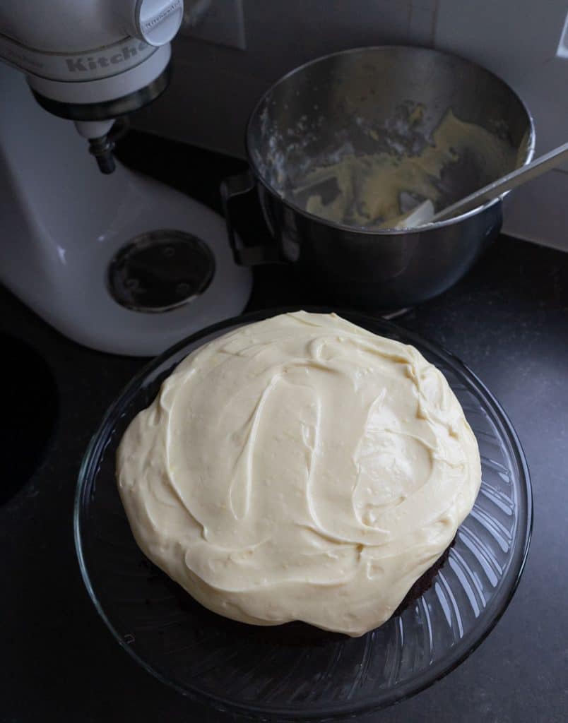 frosted best dark chocolate guinness cake on glass cake plate with the bowl of frosting and a stand mixer in the background
