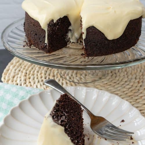 chocolate cake on a glass plate stand with slice of guinness stout chocolate cake on a white plate with a green and white gingham napkin under the plate. fork is on plate