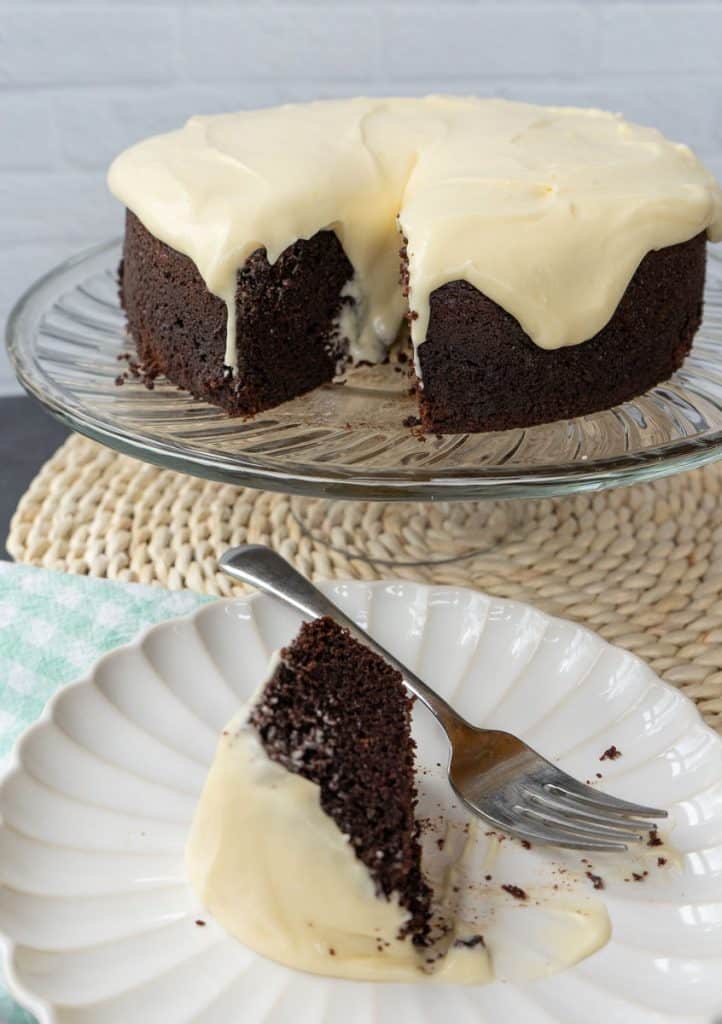 chocolate cake on a glass plate stand with slice of guinness stout chocolate cake on a white plate with a green and white gingham napkin under the plate. fork is on plate