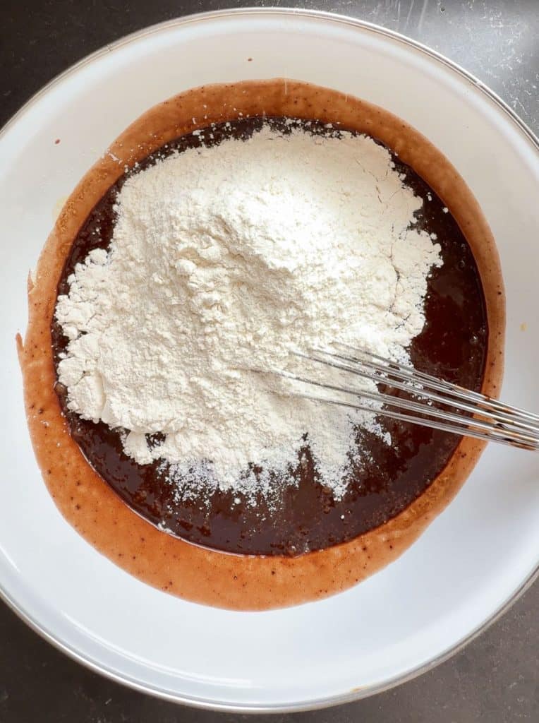 adding dry ingredients to wet ingredients in a white bowl whisk in sitting in bowl