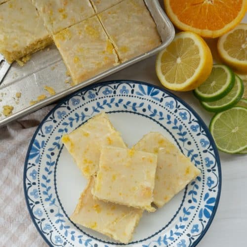 citrus bars stacked on a plate with a blue rim there are citrus fruits and the pan of citrus bars in the background