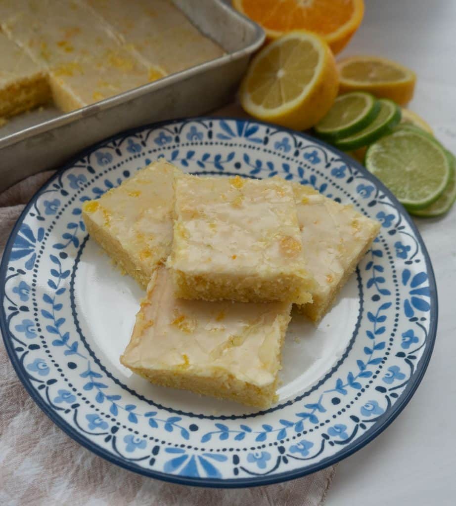 citrus bars stacked on a plate with a blue rim there are citrus fruits and the pan of citrus bars in the background