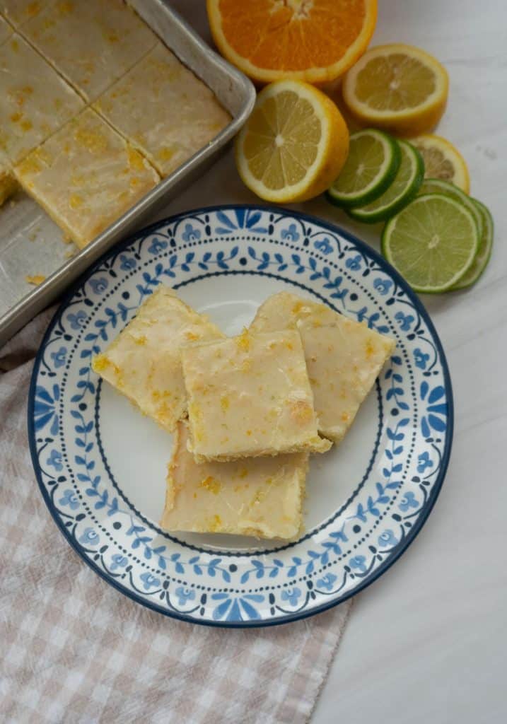 plate of citrus bars with fresh cut citrus next to it, the pan of bars is in the upper left hand corner