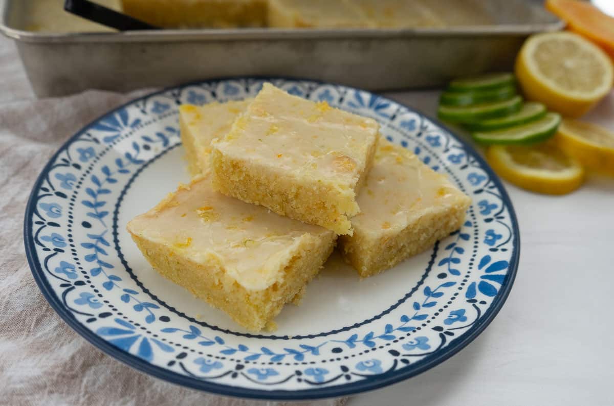 plate of glazed citrus butter bars on a blue rimmed plate with the pan of bars in the background