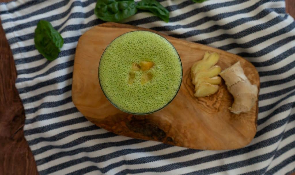 blue and white striped cloth with a wood plank on it, the green smoothie is in a glass with some pieces of mango on top 