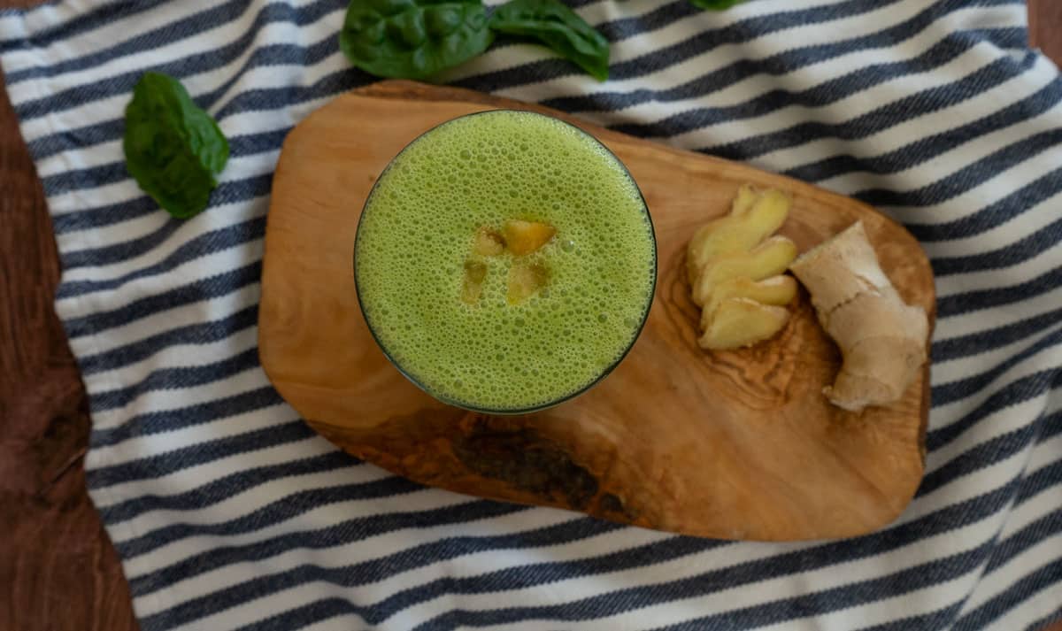 blue and white striped cloth with a wood plank on it, the green smoothie is in a glass with some pieces of mango on top