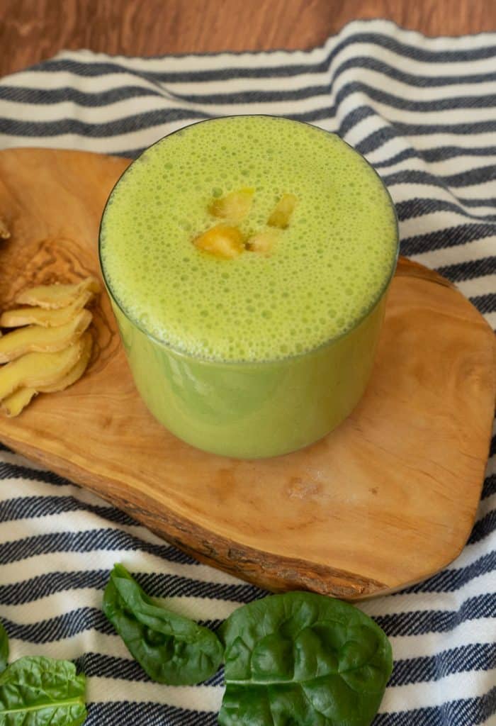 blue and white striped cloth with a wood plank on it, the green smoothie is in a glass with some pieces of mango on top