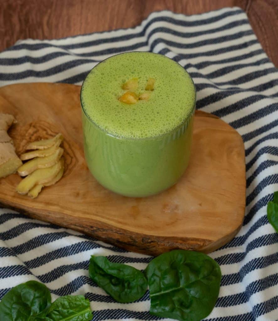 blue and white striped cloth with a wood plank on it, the green smoothie is in a glass with some pieces of mango on top