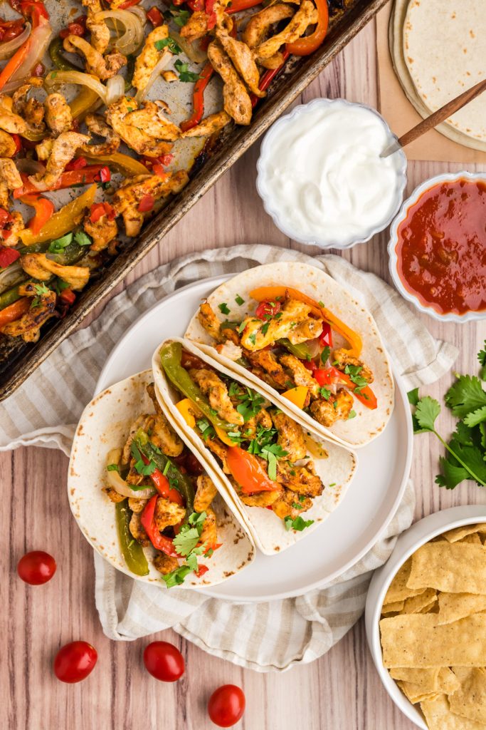 sheet pan with chicken and peppers, and three chicken fajita tacos on a plate with condiments in dishes on the side