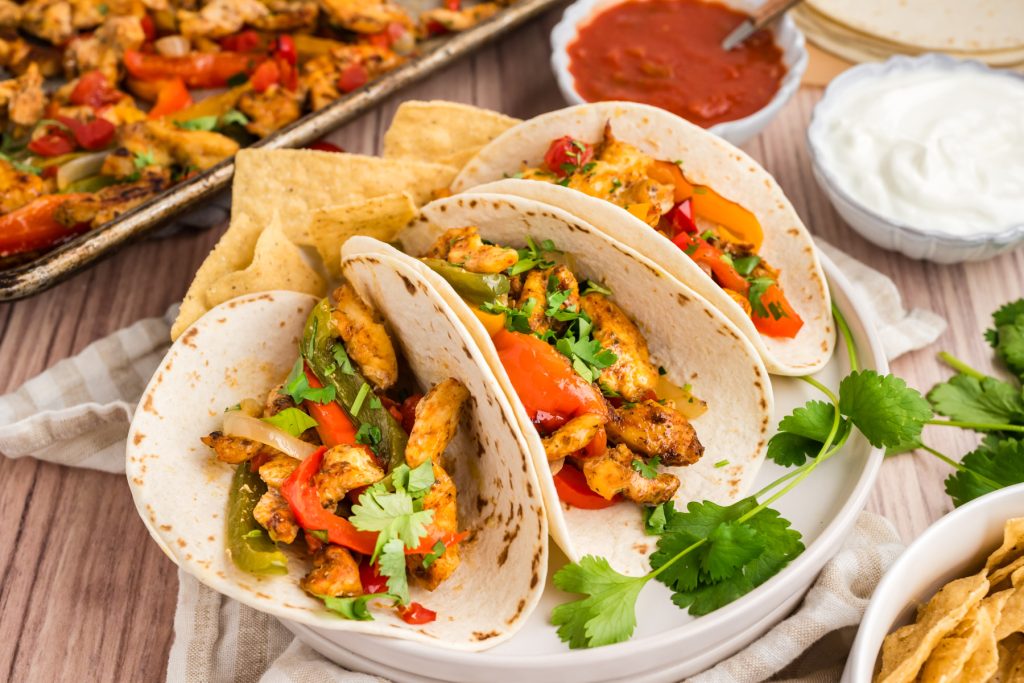 chicken and pepper fajitas in flour tortillas on a white plate with corn chips, salsa, sour cream and sheet pan fajitas behind