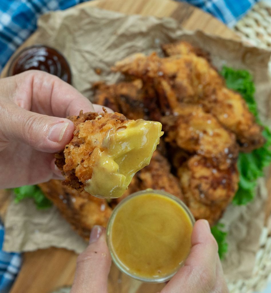 hand holding half a chicken tender that has been dipped in honey mustard sauce over a plate of chicken tenders