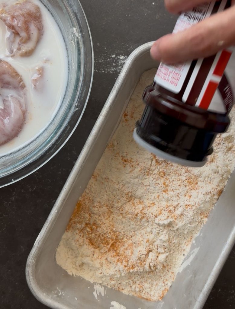 Chicken marinating in buttermilk, a pan of flour with season salt being added to it. 