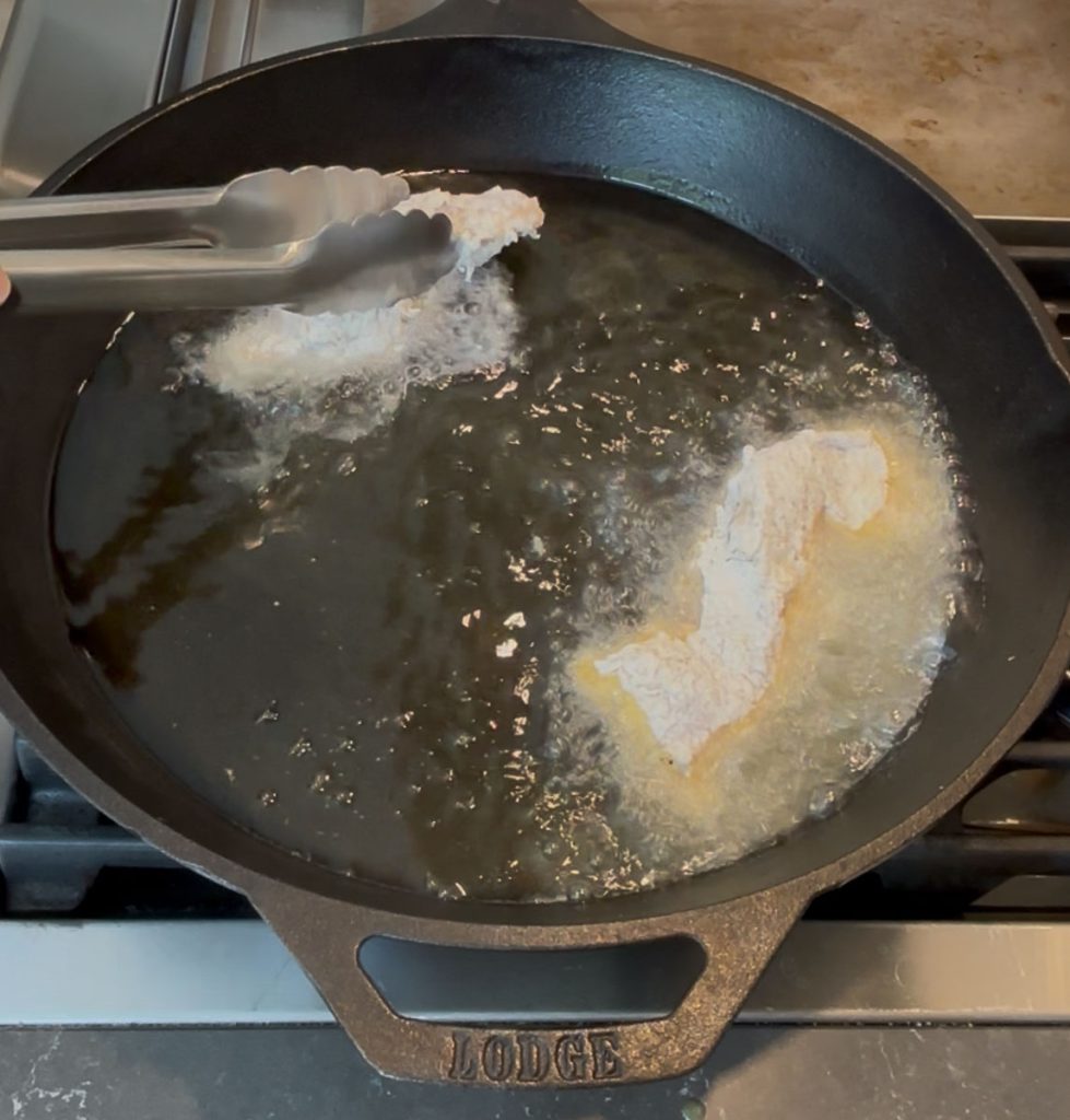 Adding battered chicken tenders to hot oil in cast iron skillet with metal tongs