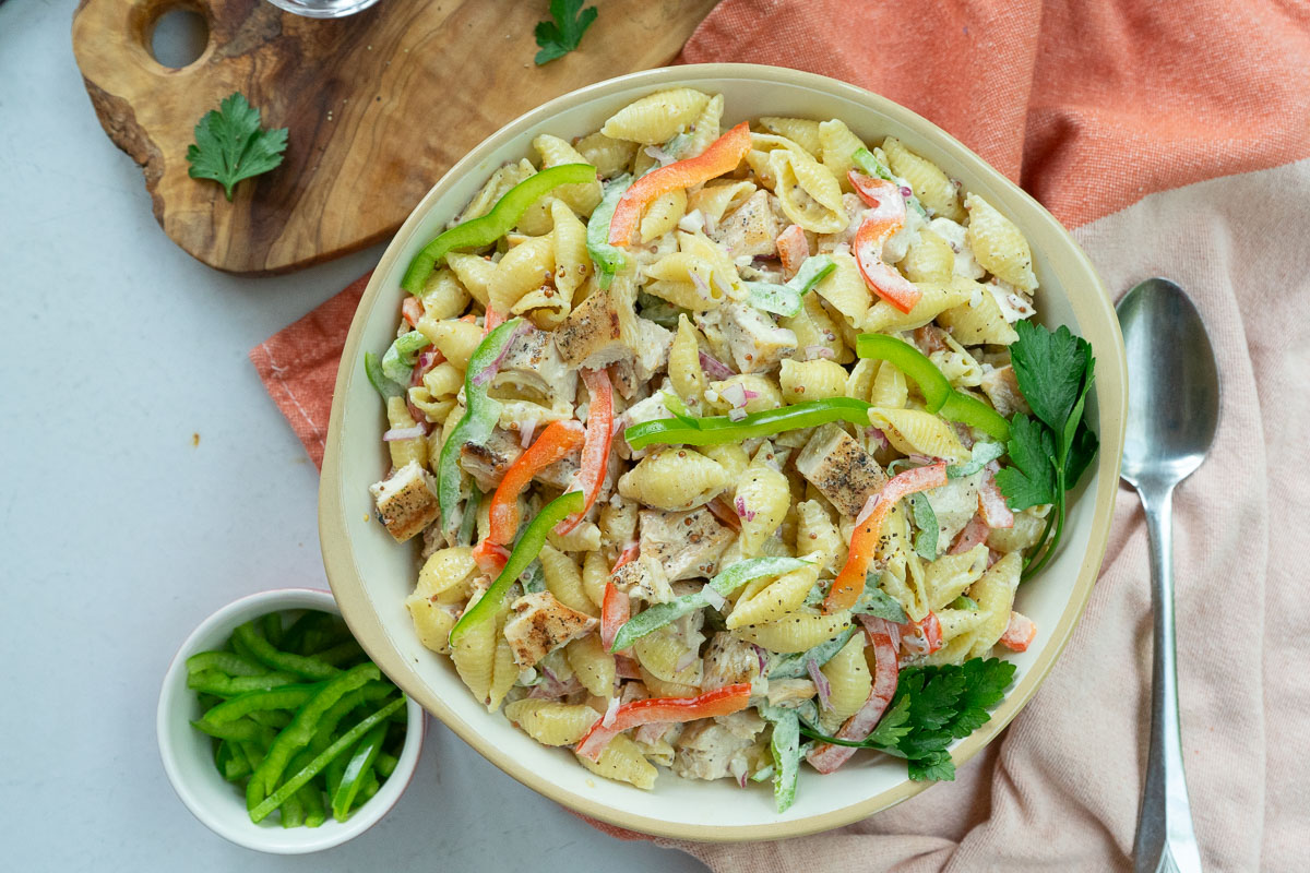 bowl of grilled chicken pasta salad with a small dish of green pepper in the lower left hand corner a peach dish towel wraps around the bottom of the bowl with a serving spoon laying on the towel