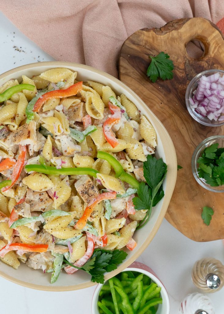 dish of grilled chicken pasta salad with a wood cutting board on the left with ingredients layed out also a serving spoon on the right with a small dish of green pepper and a peach colored dish towel
