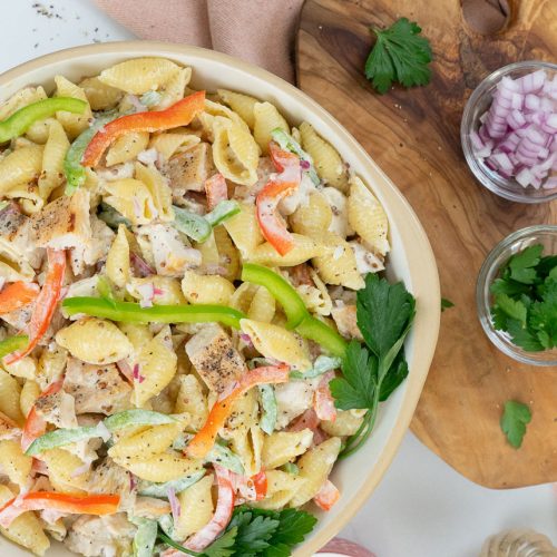 dish of grilled chicken pasta salad with a wood cutting board on the left with ingredients layed out also a serving spoon on the right with a small dish of green pepper and a peach colored dish towel