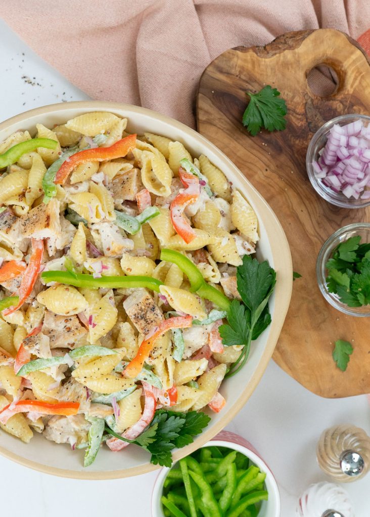 dish of grilled chicken pasta salad with a wood cutting board on the left with ingredients layed out also a serving spoon on the right with a small dish of green pepper and a peach colored dish towel