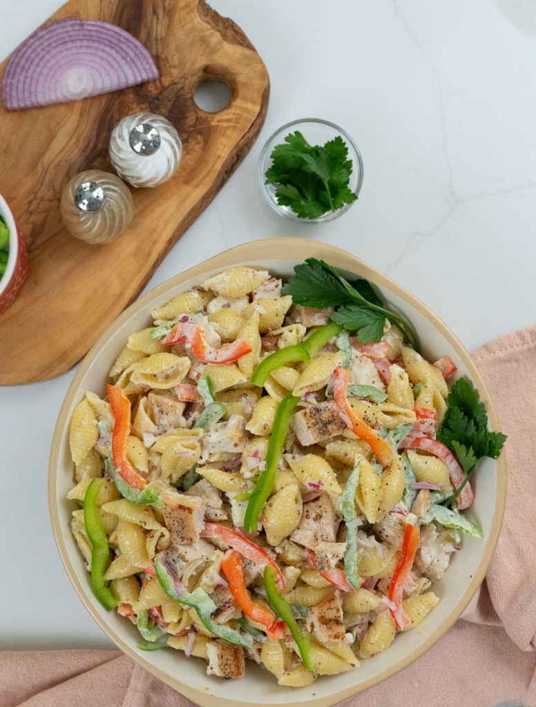 dish of grilled chicken pasta salad with a wood cutting board on the left with ingredients layed out also a serving spoon on the right with a small dish of green pepper and a peach colored dish towel