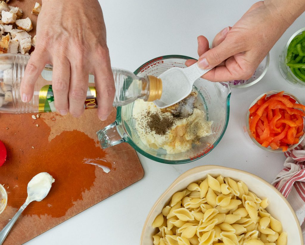 mixing pasta salad dressin in a small glass bowl with bowl of noodles in bottom of photo with peppers and other ingredients off to the right side. 