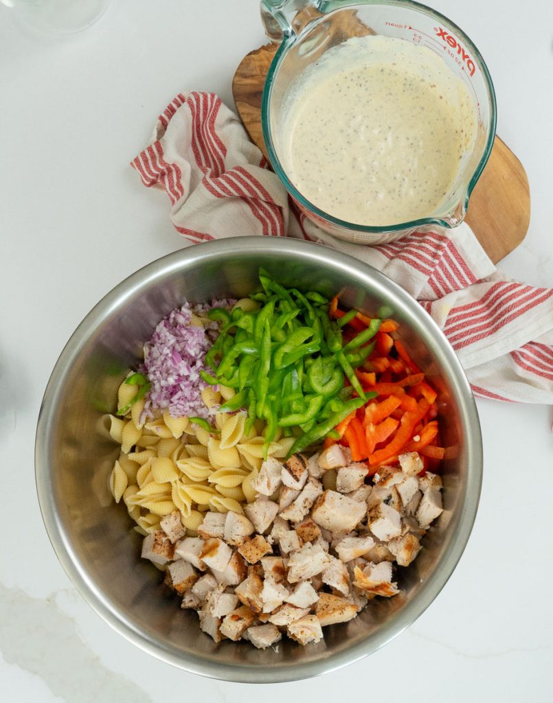 large metal bowl with pasta, chicken, onions, and peppers in it the glass bowl of salad dressing sits off to the upper left corner