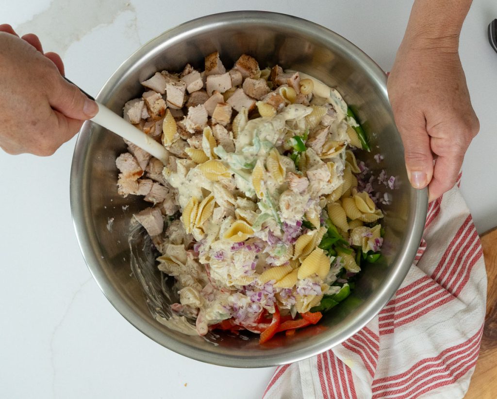 stirring pasta salad ingredients together in a large metal bowl on a white countertop with a red and white striped dish towel laying off to the right side