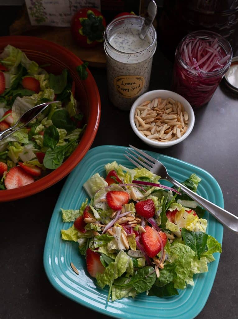 arial view of strawberry spinach salad on a light blue plate with a fork and the larger bowl salad above the plated salad with the jar of dressing and a small white bowl of toasted almonds in the upper right corner