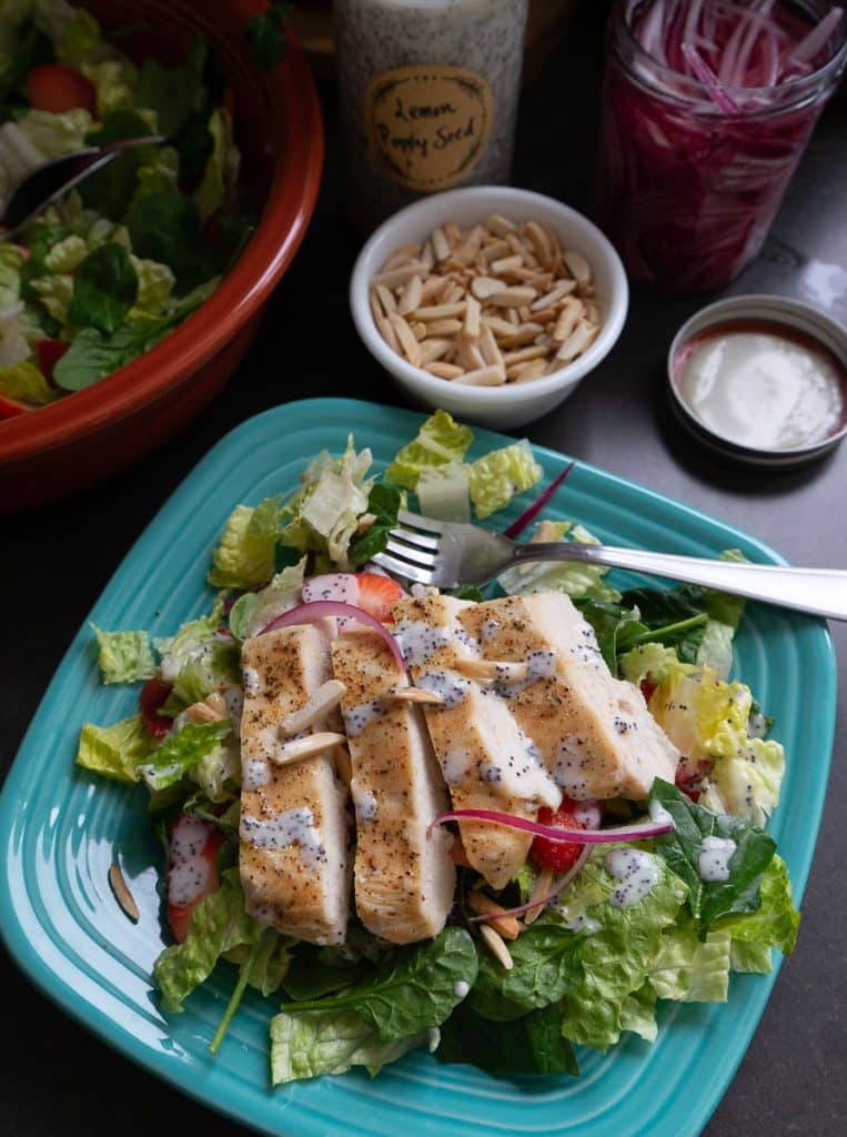 photo of a strawberry spinach salad on a light blue plate with the larger bowl of salad in the back ground with a the jar of poppy seed dressing and a small bowl of toasted almonds  with sliced chicken on top of the salad