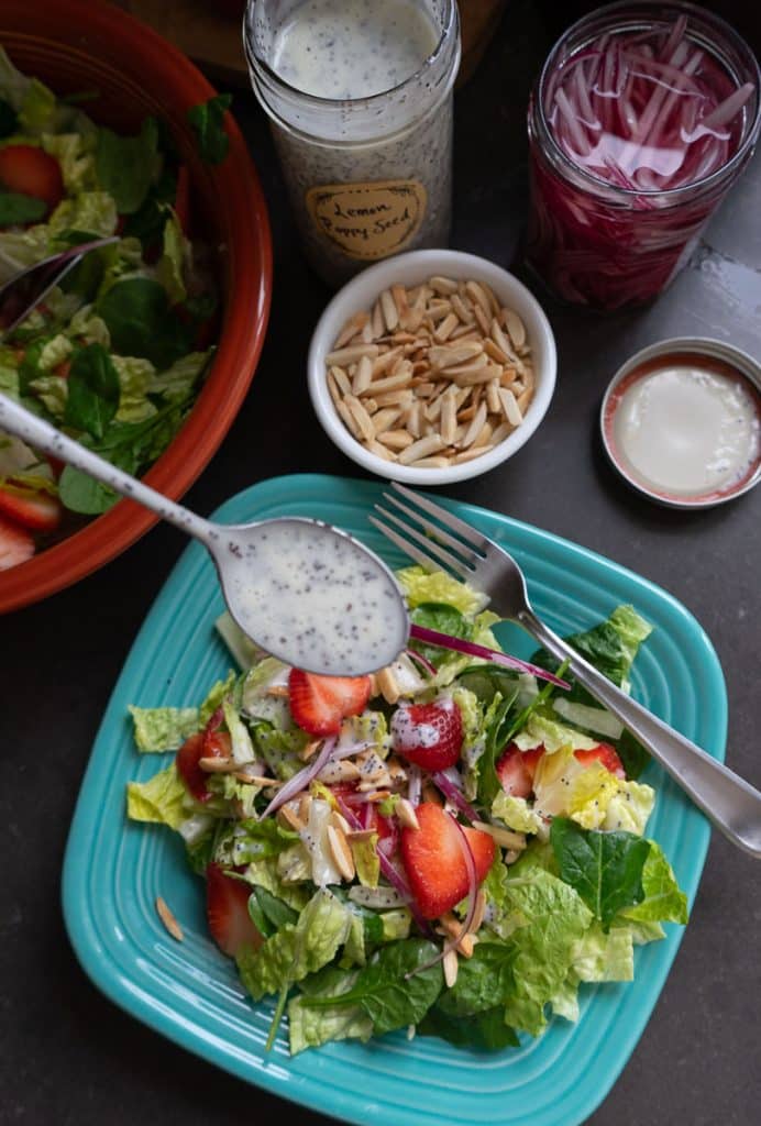 photo of a strawberry spinach salad on a light blue plate with the larger bowl of salad in the back ground with a the jar of poppy seed dressing and a small bowl of toasted almonds