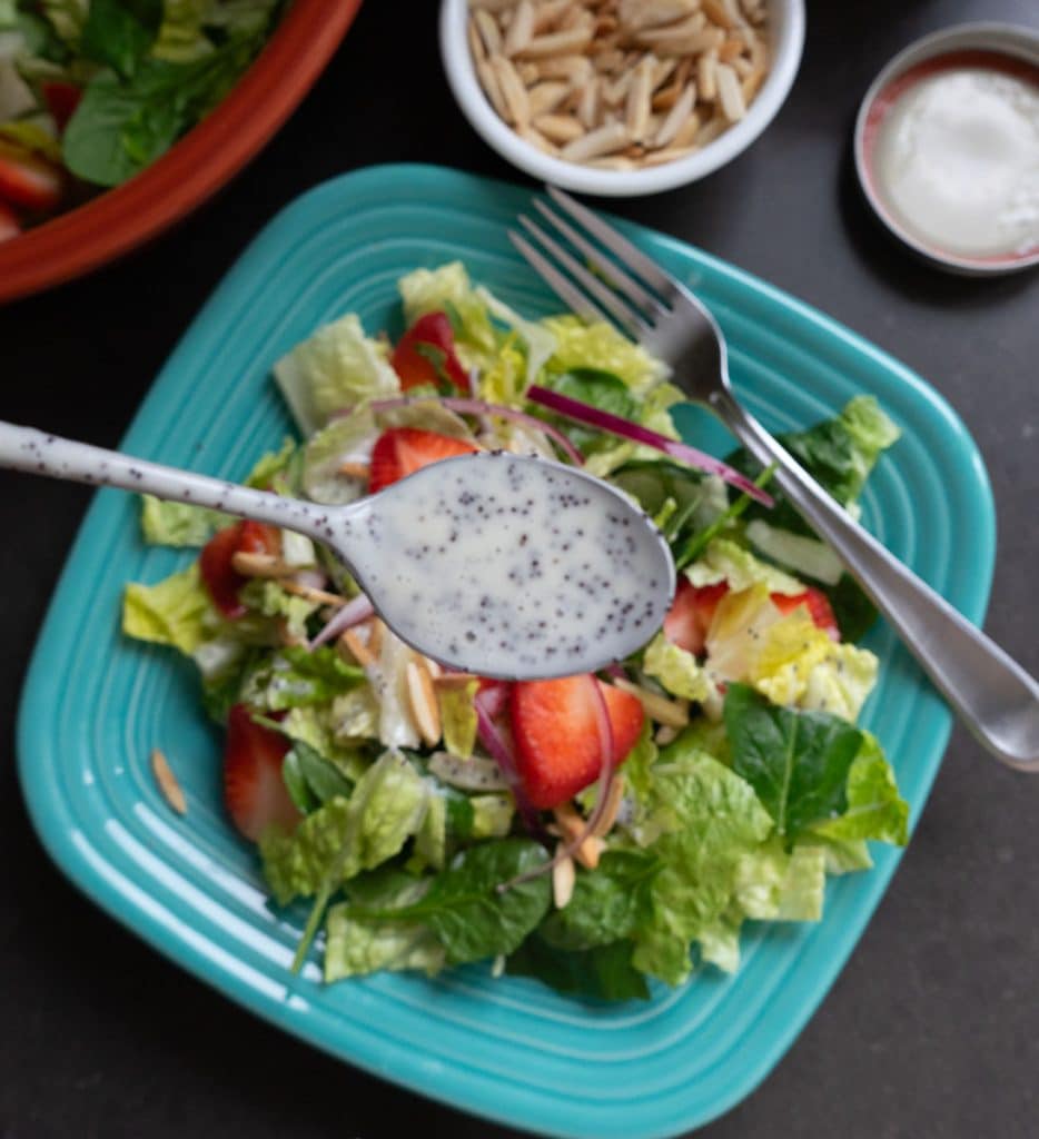 photo of a spoon drizzling poppy seed dressing over salad plate of lettuce and strawberries