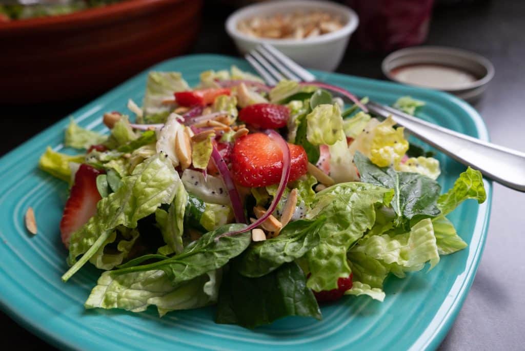 strawberry spinach salad on a light blue plate with a small white bowl of toasted almonds in the back ground and a fork on the plate