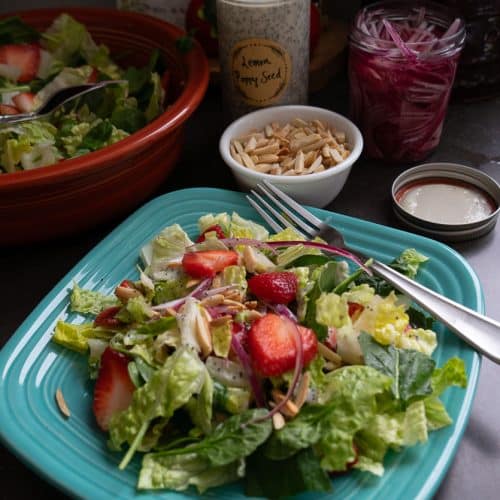photo of a strawberry spinach salad on a light blue plate with the larger bowl of salad in the back ground with a the jar of poppy seed dressing and a small bowl of toasted almonds