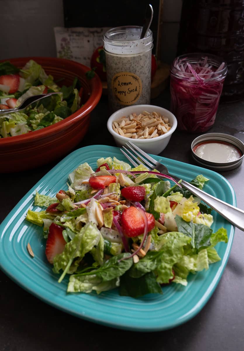 photo of a strawberry spinach salad on a light blue plate with the larger bowl of salad in the back ground with a the jar of poppy seed dressing and a small bowl of toasted almonds