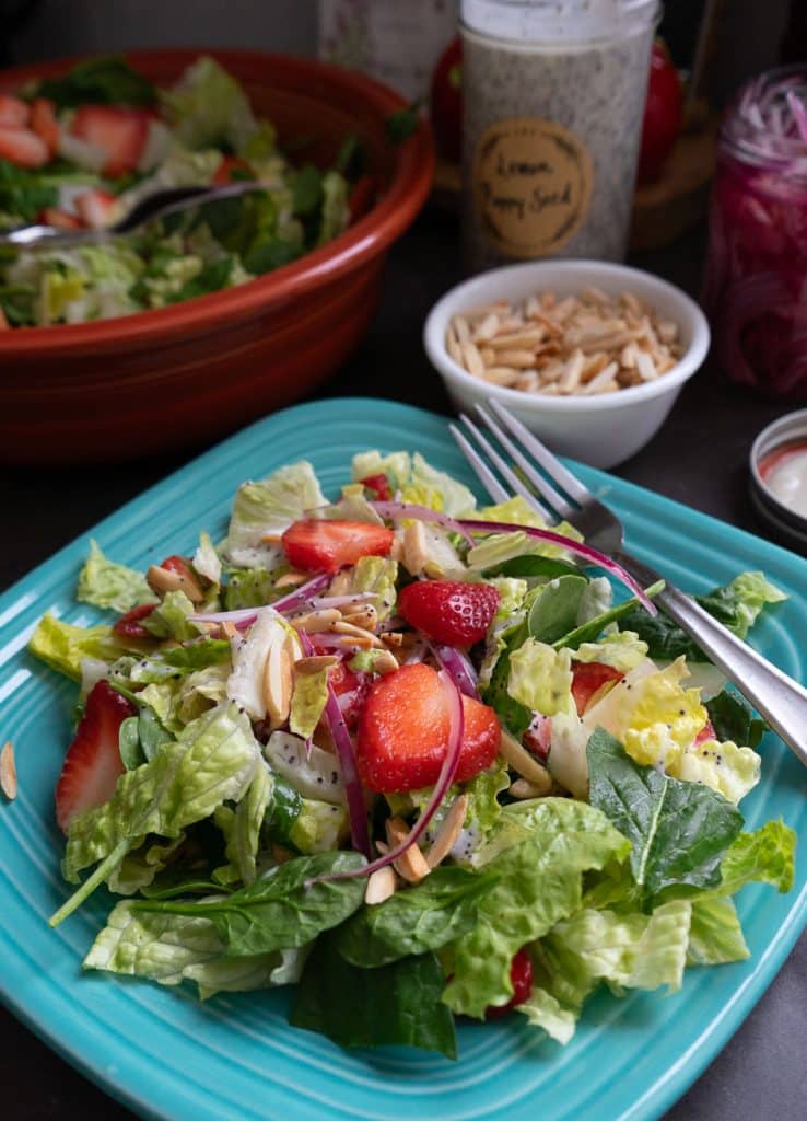 photo of a strawberry spinach salad on a light blue plate with the larger bowl of salad in the back ground with a the jar of poppy seed dressing and a small bowl of toasted almonds