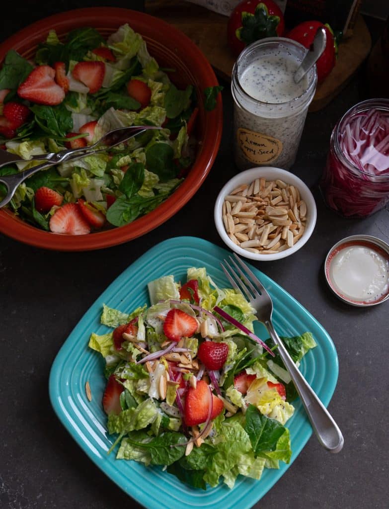 arial view of strawberry spinach salad on a light blue plate with a fork and the larger bowl salad above the plated salad with the jar of dressing and a small white bowl of toasted almonds in the upper right corner 