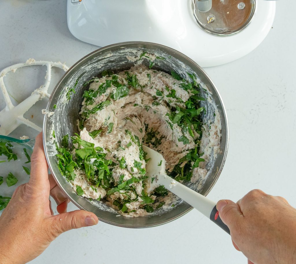 folding chopped spinach into chicken and ricotta mixture in a silver mixing bowl on a white countertop