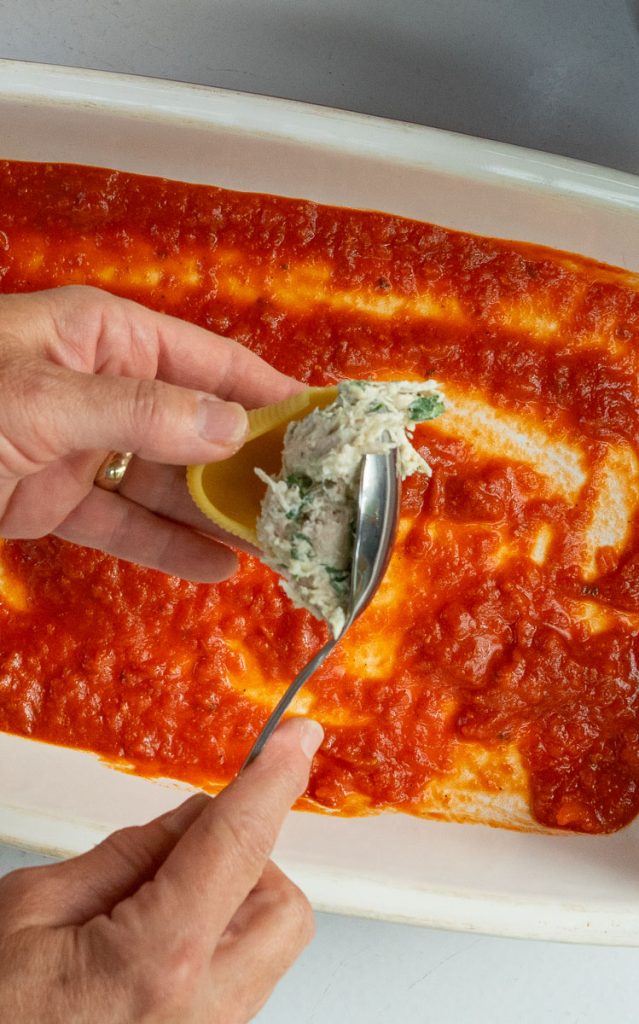 hand holding shell while other hand spoons shell with chicken ricotta mixture baking pan with red sauce in the bottom in the background