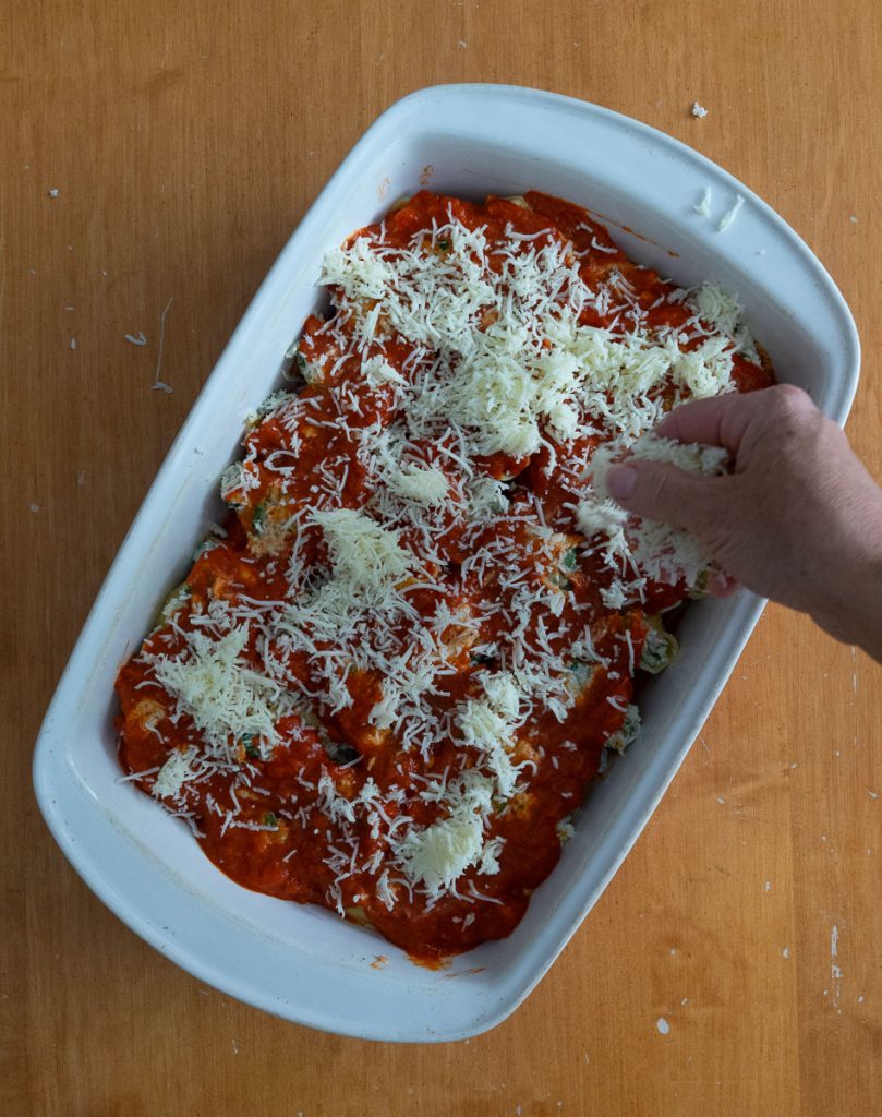 hand coming in from the right sprinkling shredded cheese on top of the pasta, dish is setting on a wood table