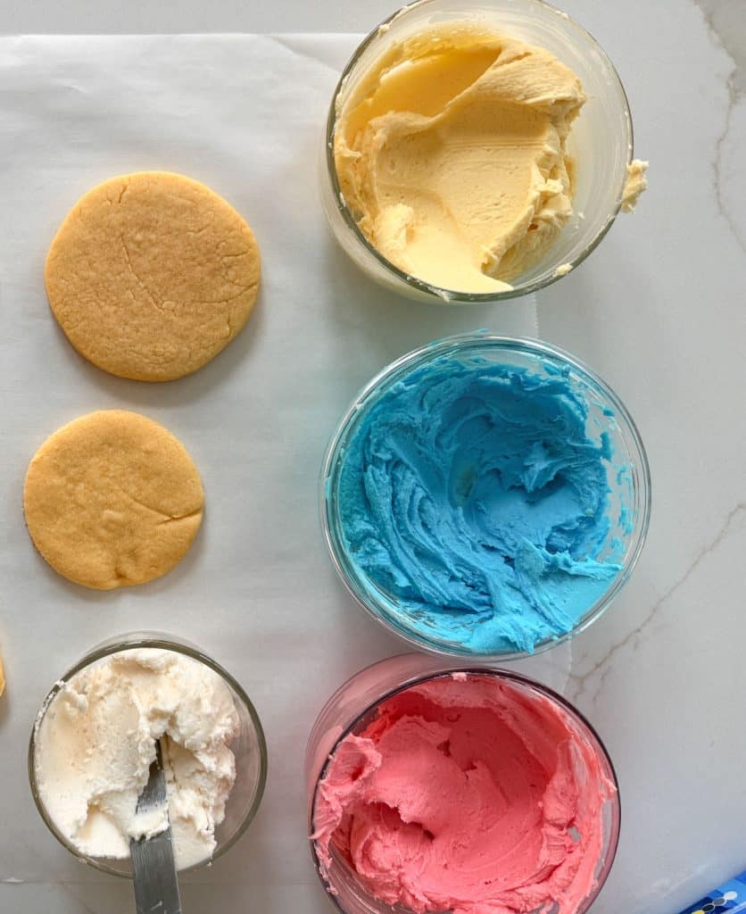 baked round cookies sitting on parchment paper with small bowls of colored frosting next to them