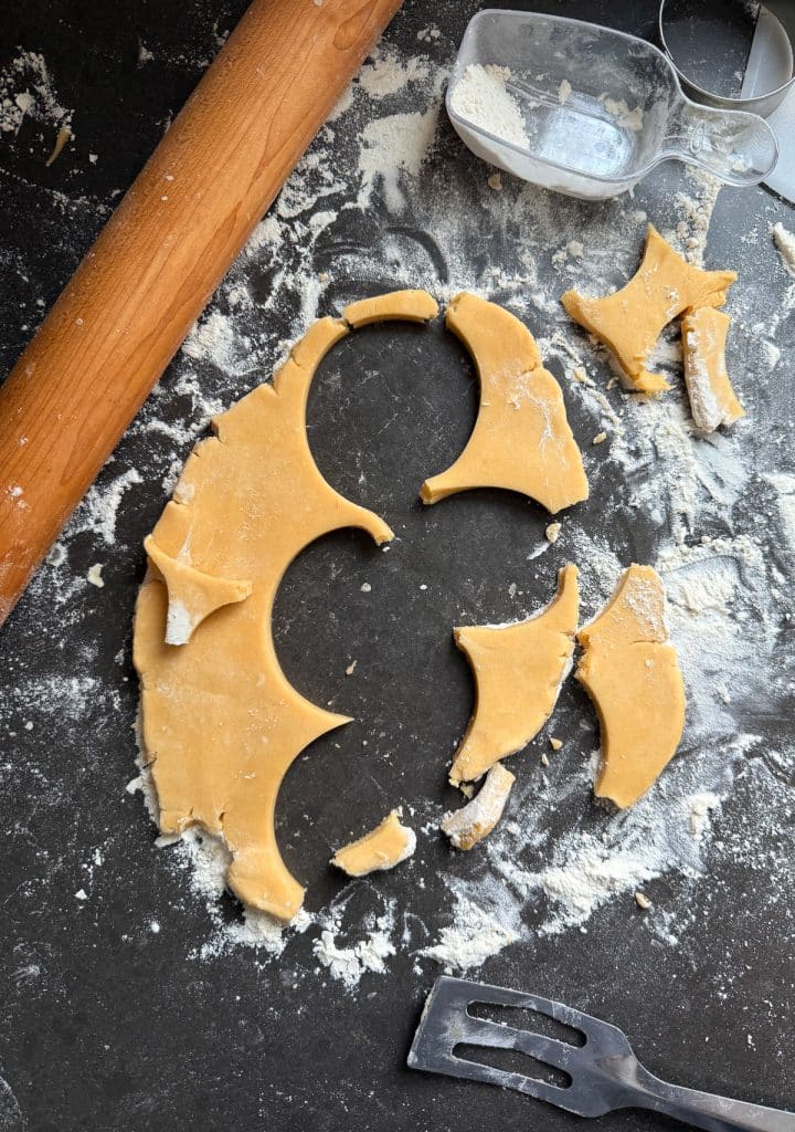 stray pieces of cookie dough after cookies have been cut out on a black countertop with a rolling pin in the upper lefthand corner