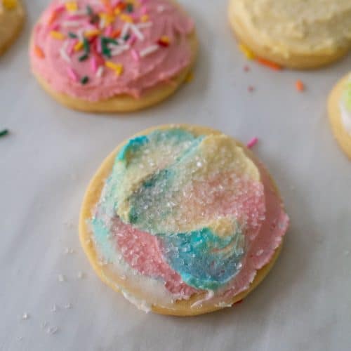 photo of a pastel frosted sugar cookie with sparkle sprinkles other frosted cookies in the background on parchment paper