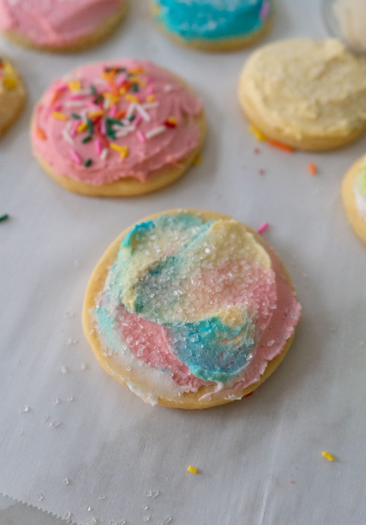 photo of a pastel frosted sugar cookie with sparkle sprinkles other frosted cookies in the background on parchment paper