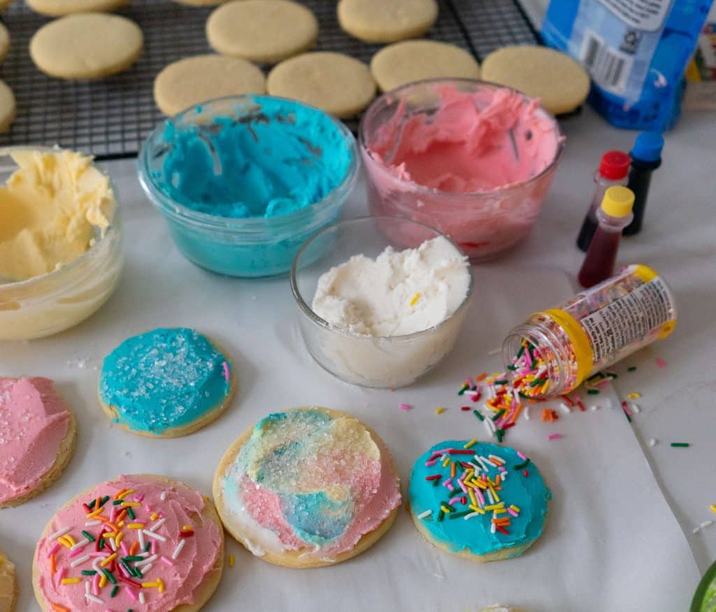 frosted cookies on parchment paper on the countertop with bowls of frosting and a bottle of sprinkles tipped over